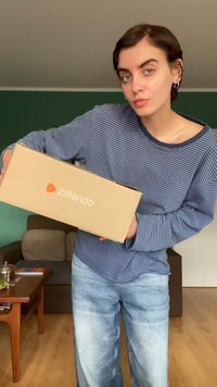 Cardboard box with "zalando" logo, held by a person. The person wears a blue and white striped long-sleeve shirt and light denim jeans.