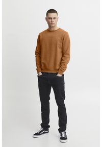 Sudadera marrón con puños y cuello acanalados, combinada con jeans oscuros y zapatillas negras. El modelo está de pie contra un fondo blanco.