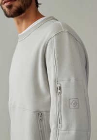 Lys grå sweatshirt med ribbet halsudskæring, side sømme og accent lynlåse på ærmerne. Har en tekstureret lap med et diamantlogo.