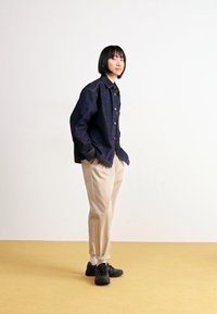 Casaco de denim azul escuro com fecho de botão, calças beijas ajustadas e sapatilhas pretas num chão amarelo com fundo branco.