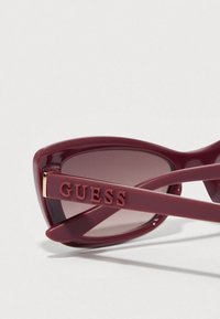 Μπλε πλαστικά γυαλιά ηλίου σε σχήμα γάτας, με φακούς με διαβαθμισμένο χρώμα και ανάγλυφη σήμανση "GUESS" στους βραχίονες. Λεπτομέρεια με χρυσή απόχρωση.