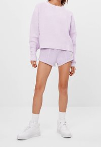 Jasnoróżowy sweter z dzianiny z długimi rękawami w komplecie z dopasowanymi fioletowymi szortami z zaokrąglonym dołem i zielonym przyciskiem z logo. Białe sneakersy.