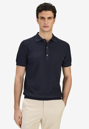 Man med kort lockigt hår bär en mörkblå kortärmad pikétröja och ljusbeiga byxor, ståendes mot en vit bakgrund.