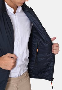 Giubbotto trapuntato blu navy con zip frontale e tasche interne. Presenta una texture liscia e dettagli a contrasto con il tirazip arancione.