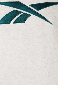 Tela gris con una superficie texturizada, con un diseño de logo bordado en teal que comprende formas angulares en la parte superior izquierda.