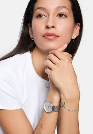 Montre-bracelet argentée avec un cadran rond et un bracelet en maille, accompagnée d'un délicat bracelet en argent. Le modèle a de longs cheveux bruns et porte une chemise blanche.