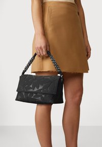 Schwarze Handtasche mit geprägtem floralen Muster, strukturiertem Material, Umklappverschluss und geflochtenem Griff, neben einem beigefarbenen Rock präsentiert.