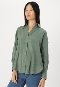 Grønn, lett rynket, langermet skjorte med krage, med hvite knapper og løs passform. Båret med blå jeans.