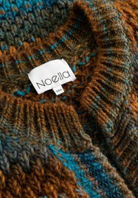 Nahaufnahme eines dicken, gestrickten Pullovers mit braunem, türkisfarbenem und blauem Garn und einem weißen Kleidungslabel mit der Aufschrift "Noella S/M", das am Kragen angenäht ist.