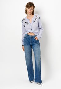 Lichtpaarse cropped cardigan met vergulde bloemenversieringen, gecombineerd met lichtblauwe flared jeans. De outfit heeft een knoopdetail aan de voorkant.