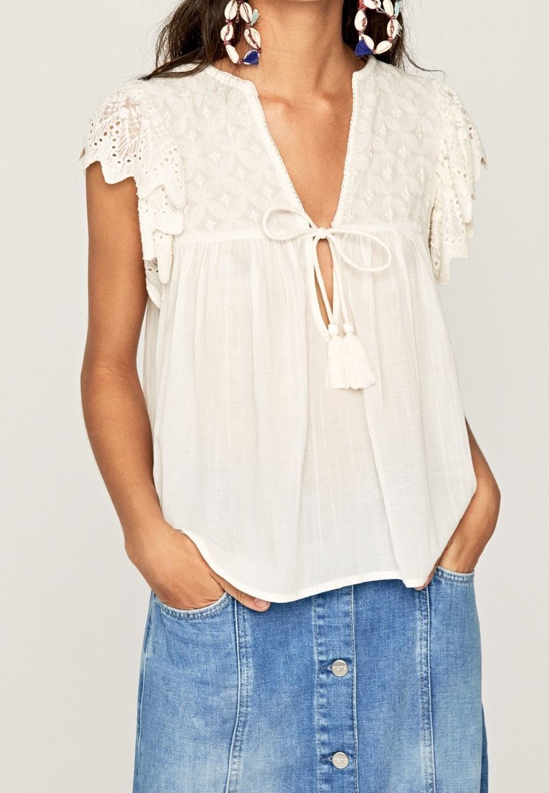 Witte, lichte blouse met scalloped, geborduurde mouwen, een V-halslijn en een geknoopte voorkant met franje. Gepaard met een denimrok.