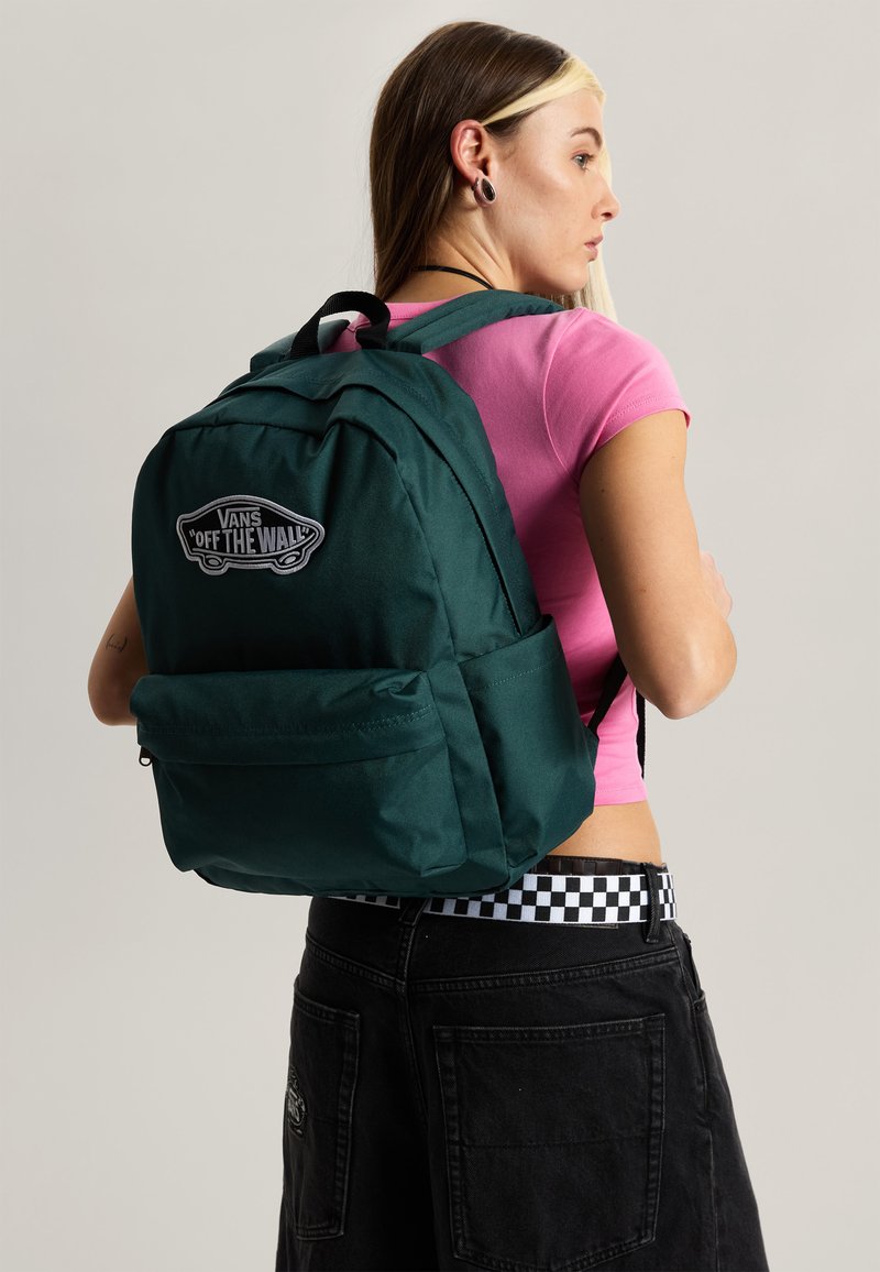 Giovane con zaino Vans verde scuro, maglietta rosa, pantaloni neri con cintura a quadri e dilatatori alle orecchie, visto di profilo.