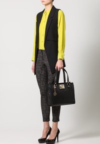 Un gilet noir sur une blouse jaune vif, associé à un pantalon à carreaux ajusté et des chaussures à talons hauts noires, tenant un sac à main noir structuré.