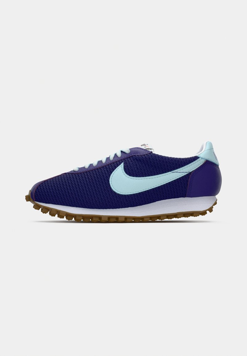 Zapatilla de malla azul oscuro con un swoosh de Nike en azul claro, cordones y pestaña del talón, media suela blanca y suela de goma natural, vista lateral sobre fondo blanco.