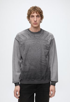Ung mand med lyst brunt bølget hår iført en grå sweatshirt med præget sol- og tekstdesign, stående mod en ensfarvet lys baggrund.