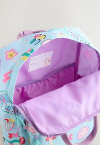 Rucksack mit einer hellblauen Außenseite, die bunte Muster aufweist. Das Innere ist lila mit einem Etikett für persönliche Angaben. Der Reißverschluss ist sichtbar.