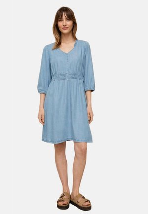 Kobieta stojąca, ubrana w jasnoniebieską sukienkę do kolan z rękawami 3/4 i beżowe sandały na platformie, na jednolitym jasnym tle.