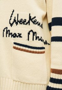 Pull en maille couleur crème avec des rayures bleu marine et marron sur la manche et la poche, avec un motif de texte en écriture bleu marine sur la poitrine.