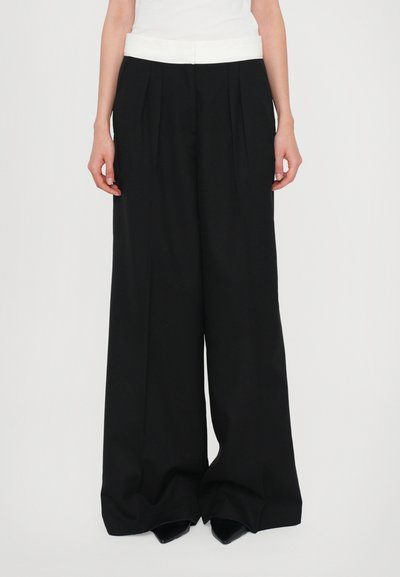 Pantalones negros de pierna ancha con una cinturilla blanca, que presentan pliegues en el frente y una textura suave, combinados con zapatos negros de punta.