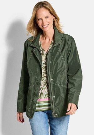 Donna sorridente che indossa una giacca verde con cerniera sopra una camicia a fantasia e jeans blu, in piedi davanti a uno sfondo chiaro e uniforme.