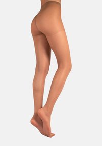 Mujer vestida con pantys nude transparentes, de pie sobre las puntas de los pies con las piernas cruzadas frente a un fondo claro.