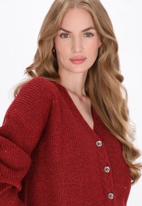 Piros kötött kardigán V-nyakkal, három fémbotrány gombbal és texturált anyaggal, kiemelve a kényelmes és elegáns kialakítást.
