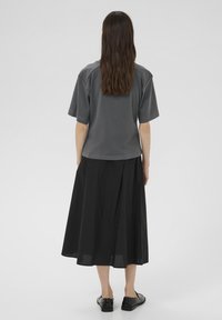 Pikkade pruunide juustega naine, kes kannab tumehallide lühikeste varrukatega särki, musta midi-seeliku ja mustade lööbastega, seisab seljaga pööratud lihtsa taustaga.