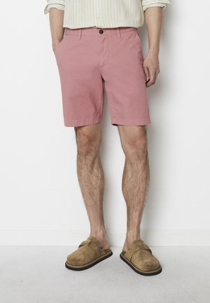 Light pink shorts made from cotton with a smooth texture, featuring a front button closure and side pockets, paired with tan suede slip-ons.
