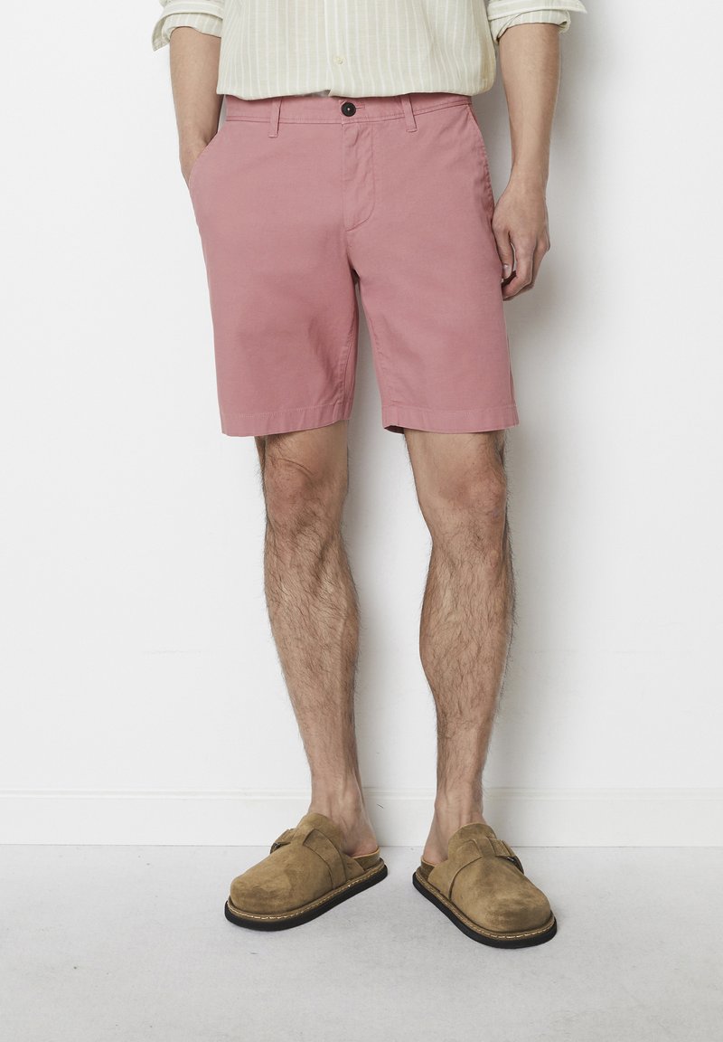 Light pink shorts made from cotton with a smooth texture, featuring a front button closure and side pockets, paired with tan suede slip-ons.