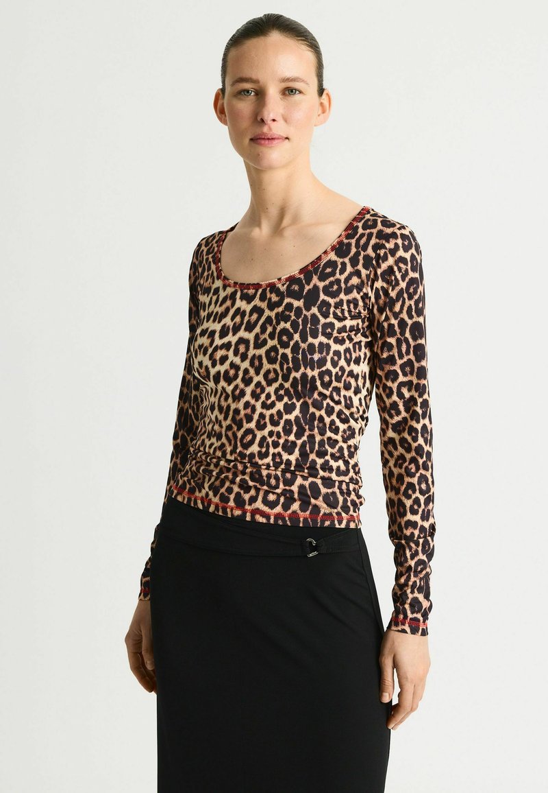 Femeie cu părul dat în spate, purtând un top cu mâneci lungi și imprimeu leopard și o fustă neagră, pe un fundal simplu, deschis la culoare.