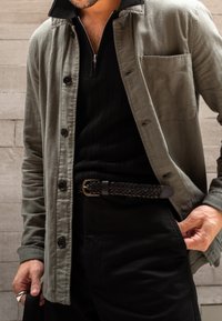 Grau karierte Hemd über einem schwarzen strukturierten Pullover, kombiniert mit dunklen Hosen und einem geflochtenen schwarzen Ledergürtel. Auffällige Details sind eine Brusttasche und ein Reißverschlusskragen.