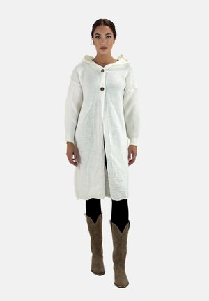 Abrigo blanco de punto con capucha, que presenta un cierre de botones y una textura acanalada. Combinado con leggings negros y botas de vaquero de color marrón.