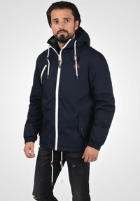 Mørkeblå jakke med hette, hvit glidelås, elastiske mansjetter og snor. Har en sidelomme og detaljer i lær.