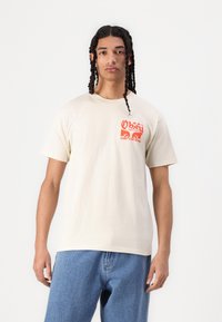 Beige bomull T-skjorte med korte ermer og rund hals. Har et rødt grafisk trykk på venstre bryst med tekst og designelementer.