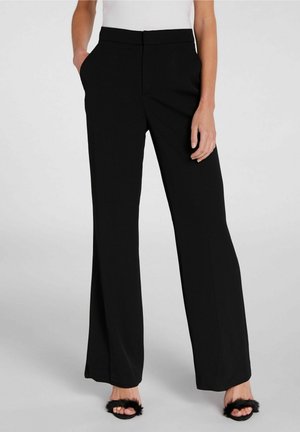 Frau trägt schwarze Hosen mit weitem Bein und Seitentaschen, kombiniert mit schwarzen offenen High Heels mit flauschigen Riemen, steht auf grauem Boden.