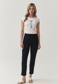 T-shirt bianca a maniche corte con design grafico blu, abbinata a pantaloni navy su misura con cintura a cravatta e sandali beige piatti.