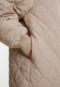 Hellbeige gesteppte Jacke mit einer Seitentasche, weichem Material und gerippten Ärmelbündchen, die Wärme und Komfort bietet.