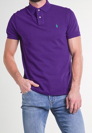 Mężczyzna ubrany w fioletową koszulkę polo z krótkim rękawem i małym zielonym logo na piersi oraz jasnoniebieskie dżinsy, stojący z jedną ręką w kieszeni.