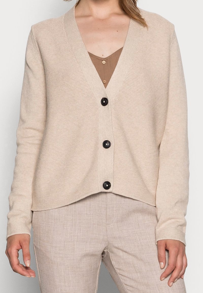 Beige gebreid vest met een V-hals, lange mouwen en een losse pasvorm, voorzien van drie zwarte knopen aan de voorkant. Glad oppervlak.
