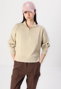 Felpa beige con zip al collo e polsini a costine. Abbinata a pantaloni cargo marrone scuro. Indossata con un cappellino da baseball rosa chiaro con logo.