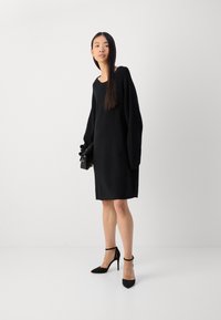 Black long-sleeve knit dress with a loose fit, featuring a rounded neckline and reaching mid-thigh. Paired with black high-heeled shoes.
