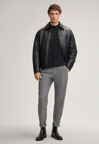 Schwarze Lederjacke mit geripptem Kragen, kombiniert mit einem strukturierten dunklen Pullover, grauen maßgeschneiderten Hosen und schwarzen Stiefeletten.