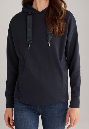 Frau trägt einen schwarzen Kapuzenpullover mit breiten Kordeln und Jeans, steht vor neutralem Hintergrund.