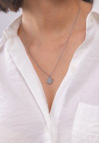 Silberne Halskette mit einem runden Anhänger, der mit einem gravierten Kompassdesign versehen ist. Die Kette ist zart und wird von einem kragenlosen weißen Hemd ergänzt.