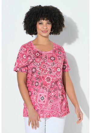 Smilende kvinde med krøllet hår iført en lyserød kortærmet skjorte med paisley-mønster og hvide bukser, stående op ad en hvid væg.