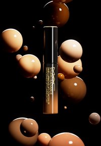 MAC Studio Radiance 24HR Luminous Lift Concealer i en klar tube med svart hette, omgitt av beige og brune sfæriske bobler på en svart bakgrunn.