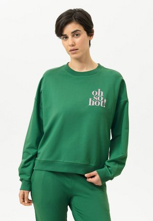 Frau mit kurzen dunklen Haaren, die ein grünes Sweatshirt und eine Hose trägt, mit dem in Pink auf der Brust gedruckten Text "oh so hot!".