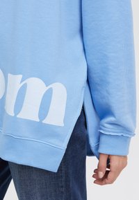 Light blue sweatshirt featuring a side slit, ribbed cuffs, and large white text on the back. Soft cotton fabric with a relaxed fit.
