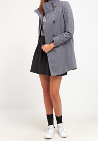 Cappotto in misto lana grigio con colletto alto, frontale doppiopetto e spalle arrotondate, abbinato a una gonna nera e sneakers bianche.