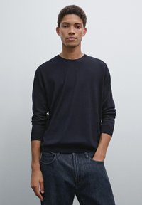 Dark navy pullover sweater with long sleeves and ribbed cuffs, paired with denim jeans. Smooth fabric with a simple round neckline.